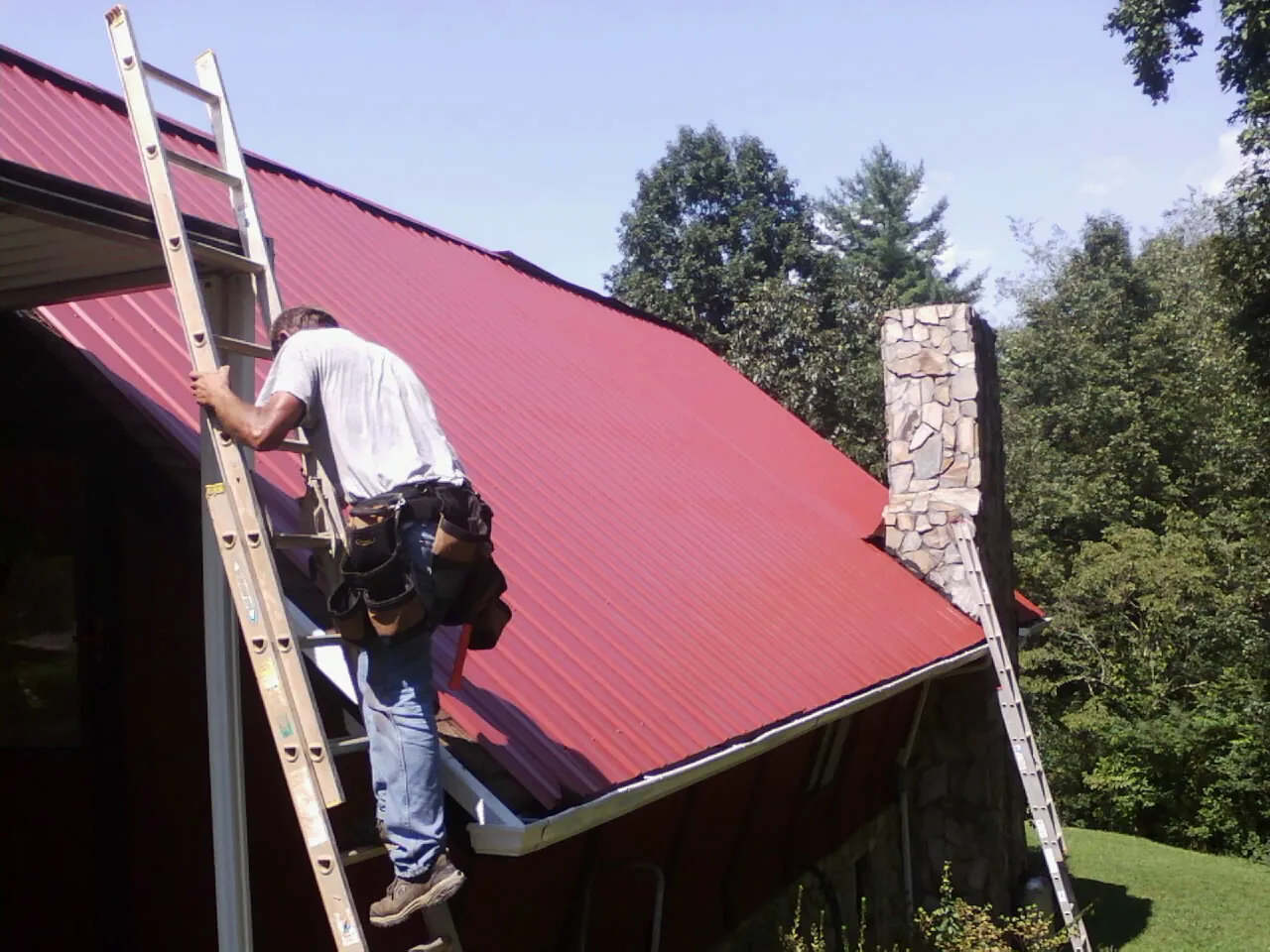 Roofer on a ladder beside a red metal roof and stone chimney — Soffit & Fascia Repair by Roofing Brothers in Sutter Creek